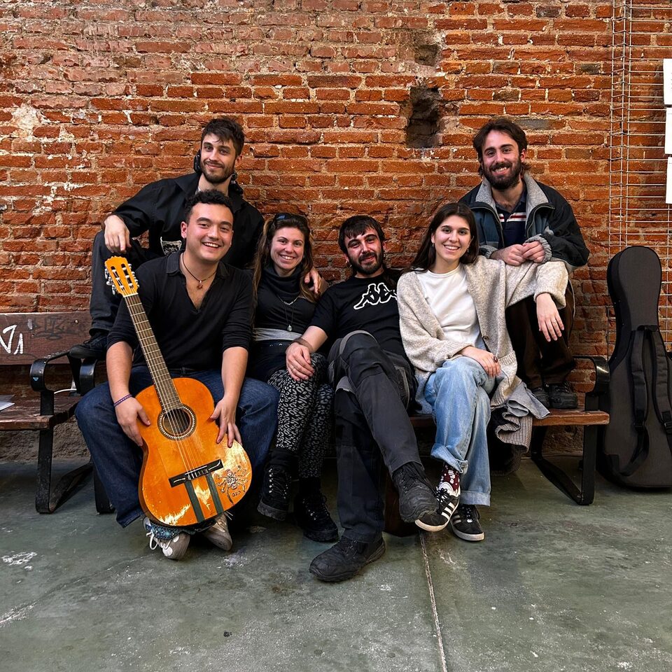 El grupo sentado en un banco ante un muro de ladrillo, con la guitarra naranja en primer plano