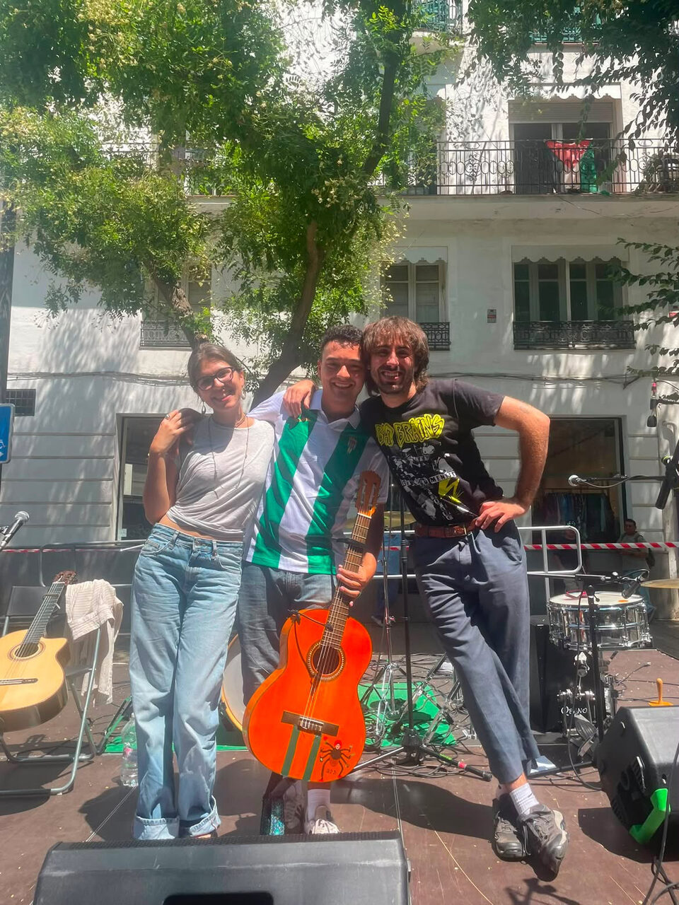 Tres músicos abrazados en un escenario montado en la calle, con la guitarra naranja por delante