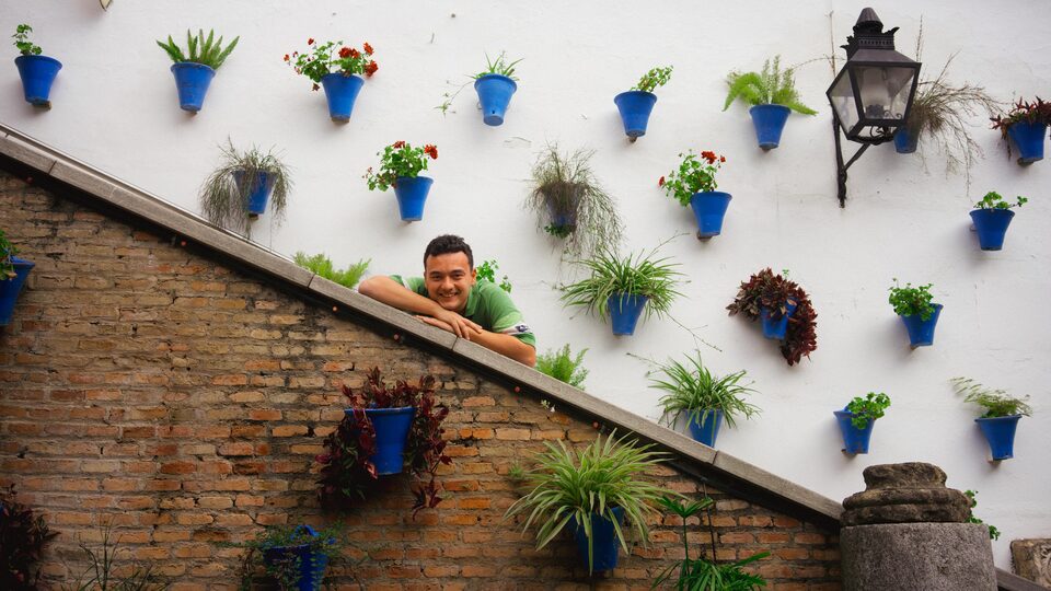 David Muntané asomado al pretil de una escalera en un patio cordobés, con la pared encalada llena de macetas azules